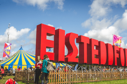 Photo showing the eisteddfod site on a bright day with clear blue skies. The huge eisteddfod letters are in he foreground with the main pavillion in the background.