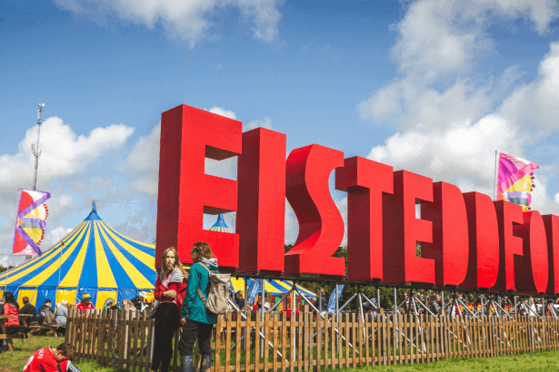 Photo showing the eisteddfod site on a bright day with clear blue skies. The huge eisteddfod letters are in he foreground with the main pavillion in the background.