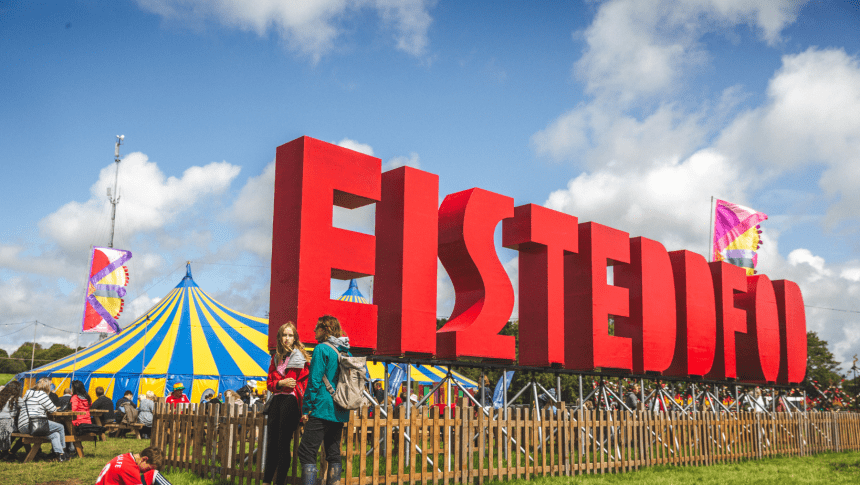 Photo showing the eisteddfod site on a bright day with clear blue skies. The huge eisteddfod letters are in he foreground with the main pavillion in the background.