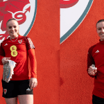 Wales Women players, Lily Woodham and Gemma Evans standing in front ofa red background withe FAW dragon logo on it, holidng the boots and the tooth they have donated to the museum.