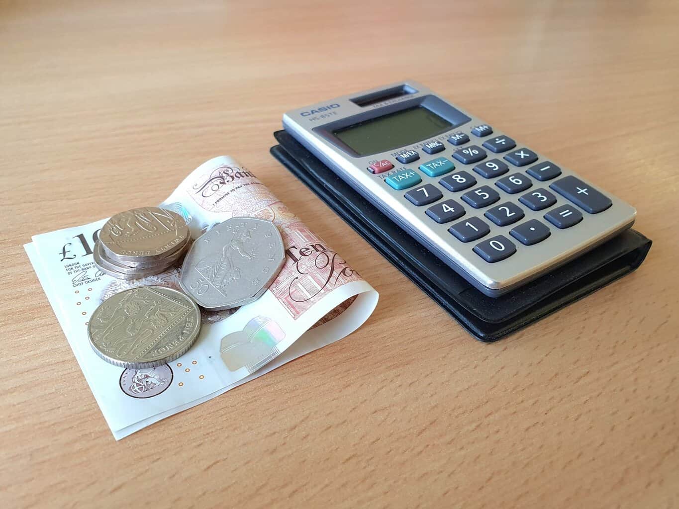 Pentwr bach o geiniogau arian ar ben nodyn £10 wedi'i blygu wrth ymyl cyfrifiannell ar ddesg